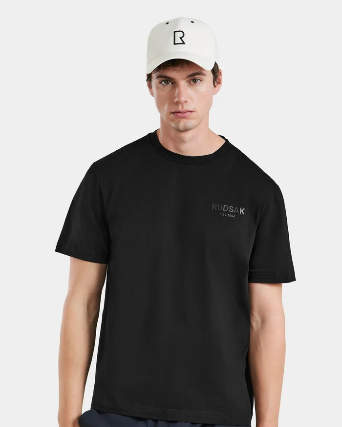 A young man wears the Rudsak ARSENE - 9422504 Iceland black t-shirt and a white unisex baseball cap with a black R logo, standing against a plain light background.
