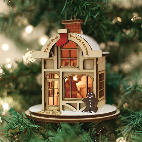 A wooden Christmas tree ornament shaped like a cozy shop, labeled Big Red Sock Stocking Company, hangs on a tree. Warm light glows from inside, and a gingerbread figure stands at the entrance.