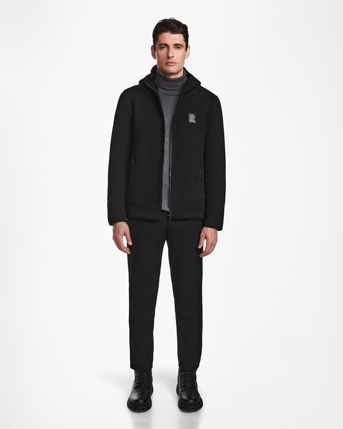 A man stands facing forward against a plain white background, wearing the Rudsak BILLY - U524501 Black M unisex zip-up sweater over a gray turtleneck, black pants, and black boots.