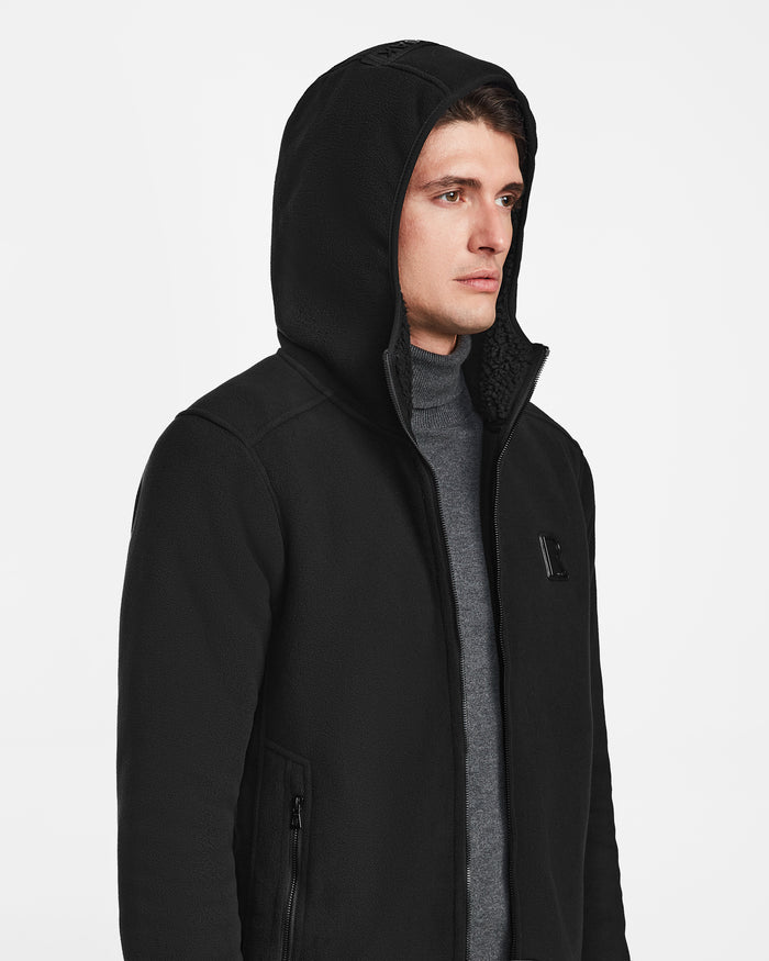 A man models the Rudsak BILLY - U524501 black polar fleece hoodie in size M over a gray turtleneck, standing against a plain white background and facing slightly to the side.