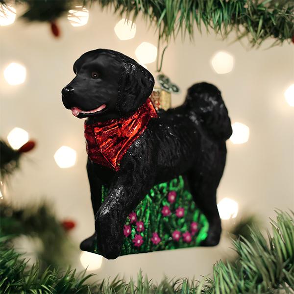 A Christmas tree ornament shaped like a black dog wearing a red scarf and standing on a green base with pink flowers, surrounded by tree branches and blurred lights in the background.