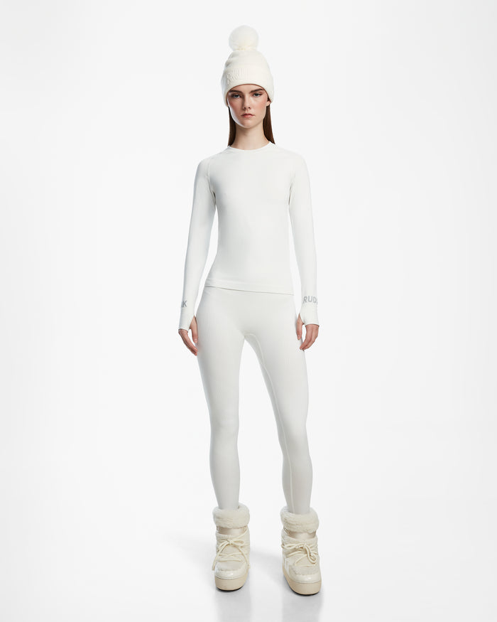 A woman faces forward against a plain white background, wearing Rudsaks CARRIE - 8524619 Iceland tall, fluffy winter boots with an all-white fitted top, stretch leggings, and a knit pom-pom beanie.