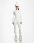 A woman faces forward against a plain white background, wearing Rudsaks CARRIE - 8524619 Iceland tall, fluffy winter boots with an all-white fitted top, stretch leggings, and a knit pom-pom beanie.