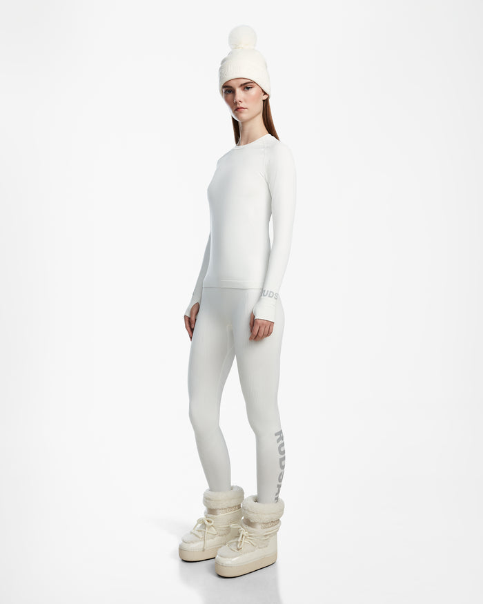 A woman wears Rudsak’s CARRIE - 8524619 Iceland: a fitted white long-sleeve top and stretch leggings ski base layer, paired with a white knit beanie with pompom and off-white winter boots, standing against a white background.