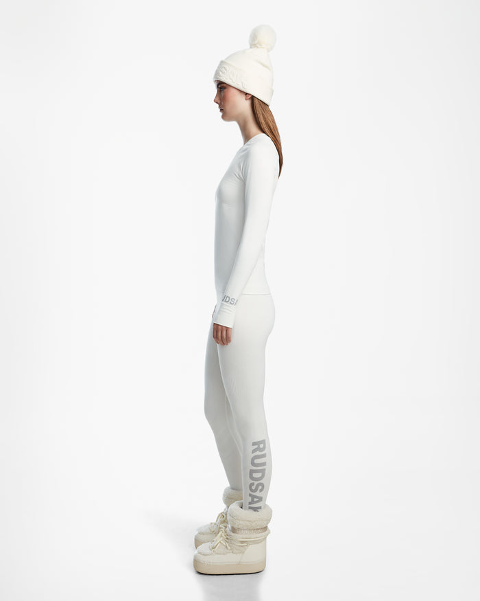 A woman stands in profile against a white background, wearing the Rudsak CARRIE - 8524619 Iceland knit hat with pom-pom, a long-sleeve women’s thermal top, stretch leggings with RUDSAK branding, and chunky white boots.