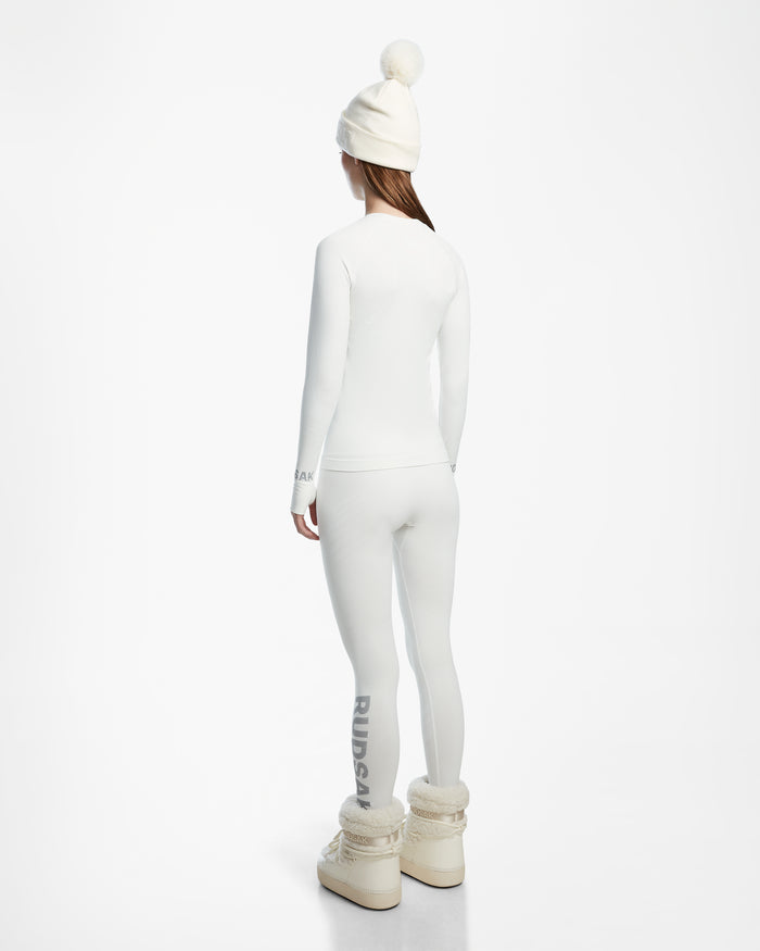 A person faces away, wearing the Rudsak CARRIE - 8524619 Iceland white ski base layer top and matching stretch leggings featuring RUDSAK text on one leg, white winter boots, and a white pom-pom knit hat against a plain white background.