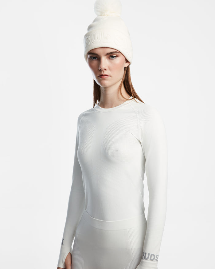A woman wearing the Rudsak CARRIE - 8524619 Iceland fitted white ski base layer and a white knit pom-pom beanie stands against a white background, looking at the camera with a neutral expression.