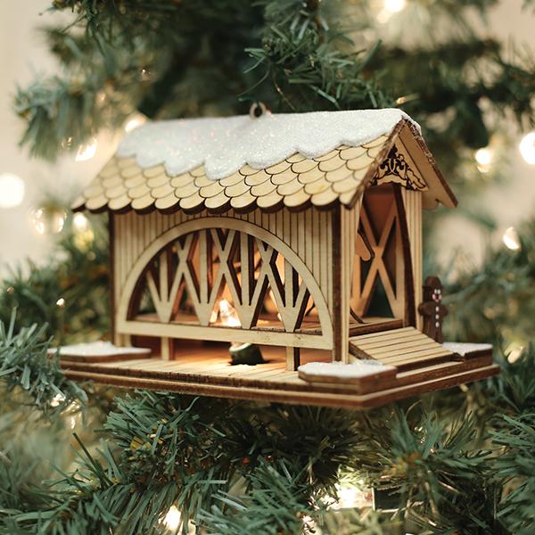 A wooden covered bridge ornament with a snow-dusted roof hangs on a Christmas tree, with soft white lights and green pine needles in the background.
