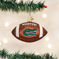 A football-shaped Christmas ornament featuring the Florida Gators logo hangs from a decorated tree branch with green needles and white lights in the background.