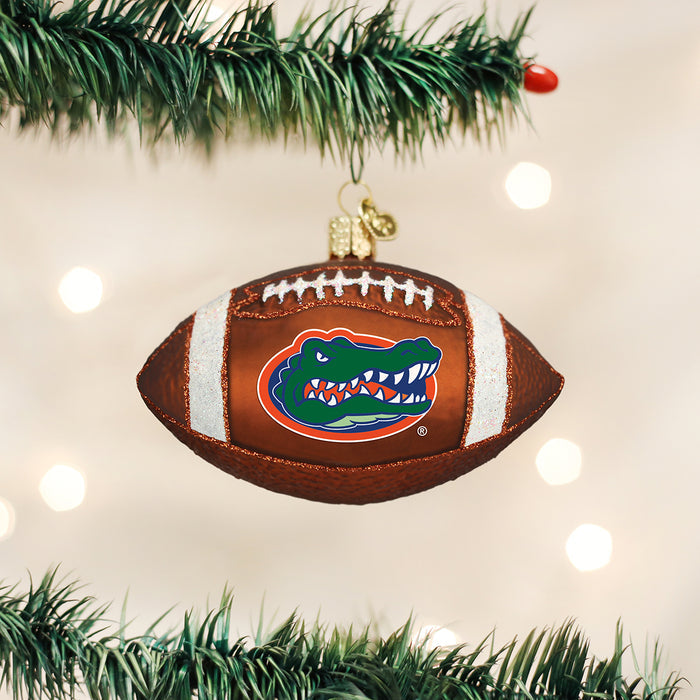 A football-shaped Christmas ornament featuring the Florida Gators logo hangs from a decorated tree branch with green needles and white lights in the background.