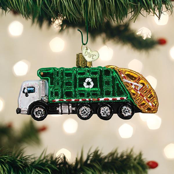 A green garbage truck Christmas ornament with a recycling symbol hangs on a tree branch. The background features blurred holiday lights.