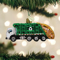 A green garbage truck Christmas ornament with a recycling symbol hangs on a tree branch. The background features blurred holiday lights.