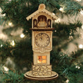 A wooden cuckoo clock ornament with Roman numerals hangs on a Christmas tree, decorated with small carved figures and a glowing window at the base. Green pine needles and lights surround the ornament.