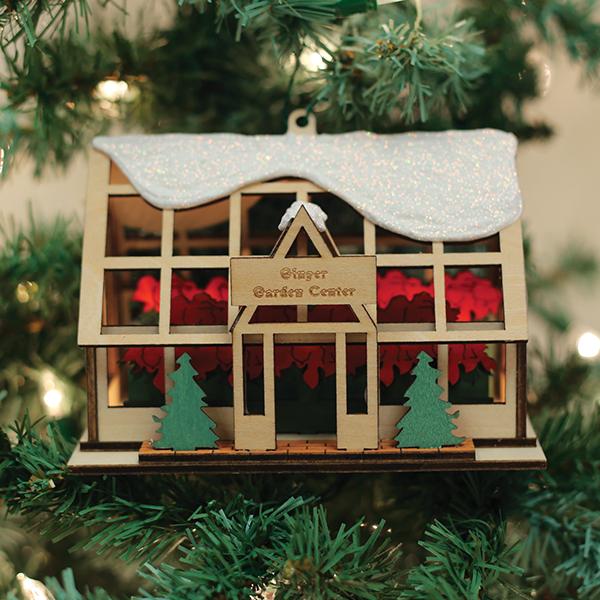 A wooden Christmas ornament shaped like a greenhouse with a glittery snow-covered roof hangs on a tree. The sign reads Ginger Garden Center and two green trees decorate the front.