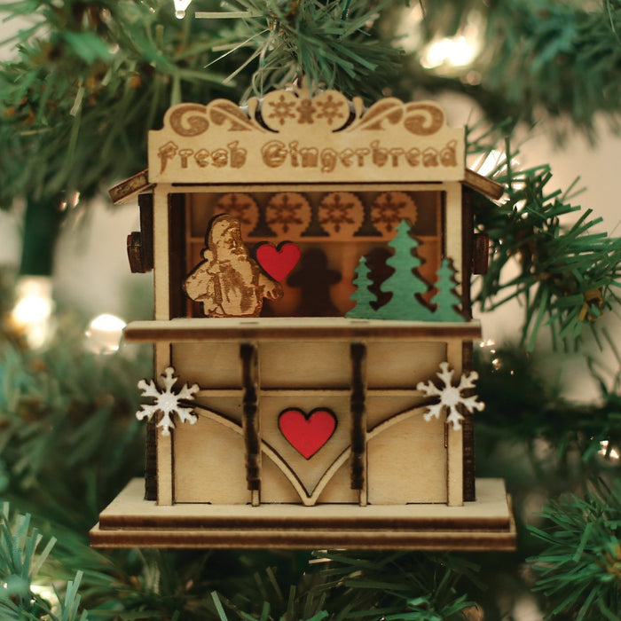 A wooden Christmas ornament shaped like a gingerbread stall hangs on a tree. The sign reads Fresh Gingerbread and features hearts, snowflakes, a gingerbread figure, and a green tree.