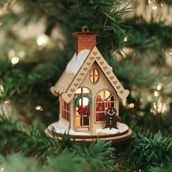 A small, cozy gingerbread house ornament with a wreath and a gingerbread figure hangs on a Christmas tree, surrounded by green branches and warm lights.