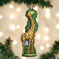 A glass Christmas tree ornament shaped like a giraffe with brown spots stands among green foliage, hanging from a tree branch, with soft holiday lights blurred in the background.