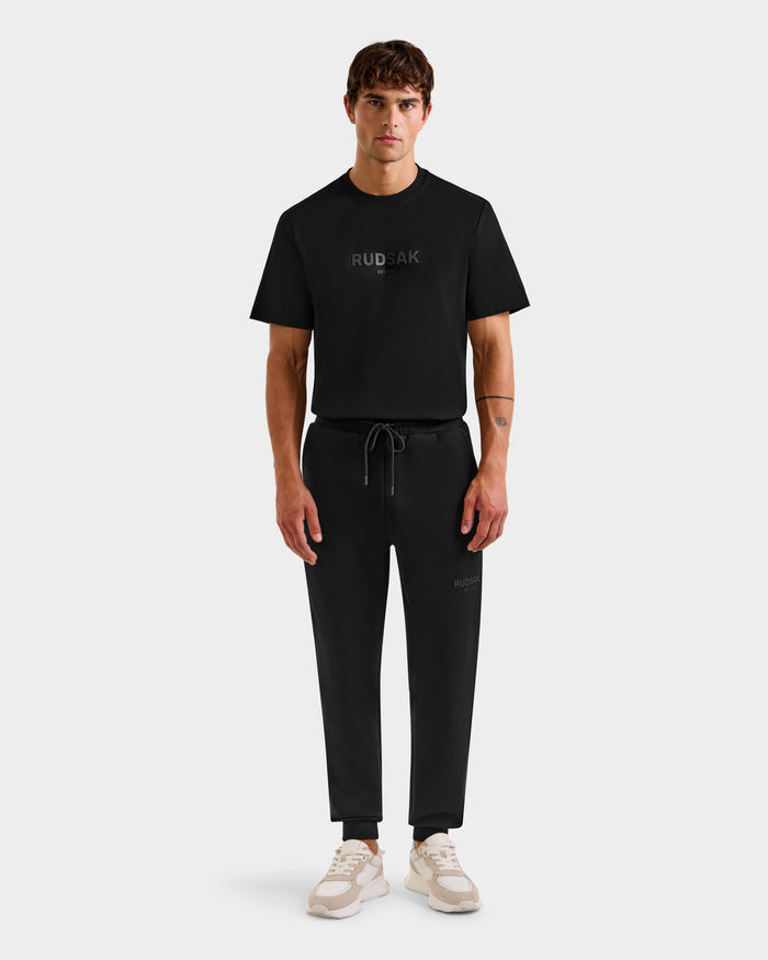 A man faces forward in Rudsaks JOSS - 9524009 Black M T-shirt and joggers, styled with beige and white sneakers. The unisex set is displayed against a plain light gray background.