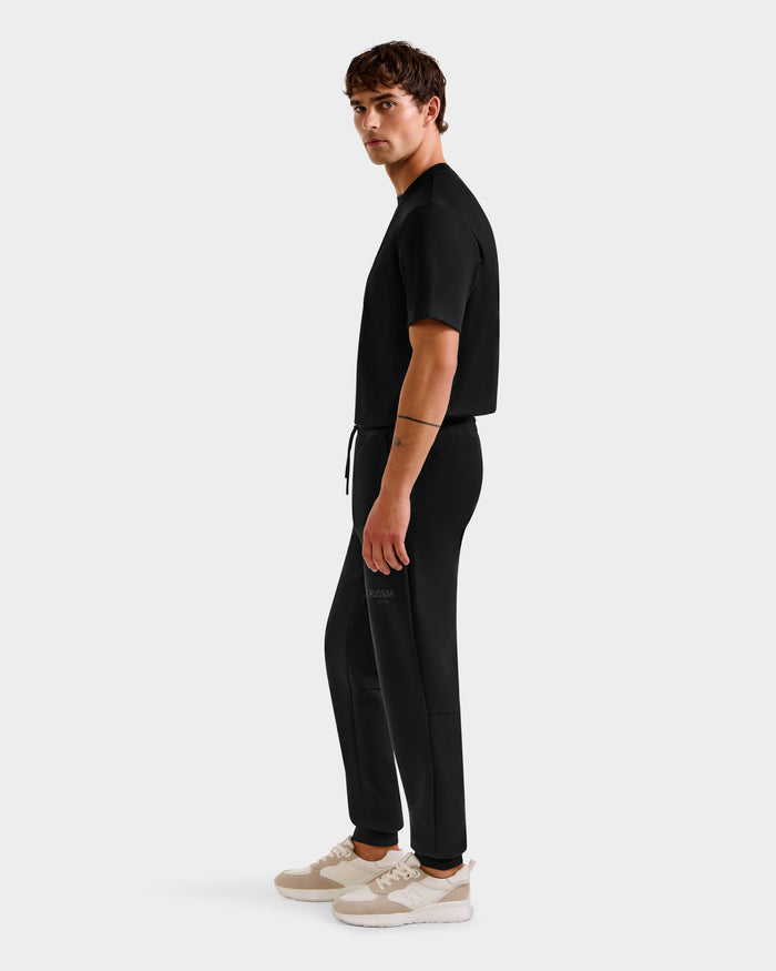 A person stands in profile against a plain background, wearing the Rudsak JOSS - 9524009 Black M short-sleeve top, stylish joggers, and beige sneakers.