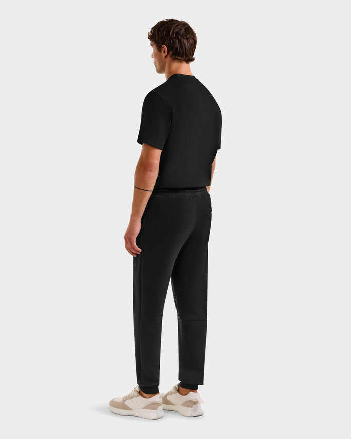 A man faces away, wearing the Rudsak JOSS - 9524009 Black M t-shirt with joggers and white sneakers against a plain light-gray background.