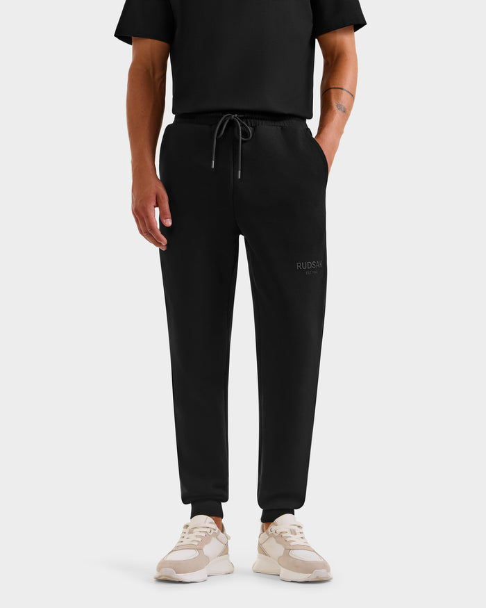 Wearing Rudsak’s JOSS - 9524009 Black M unisex joggers with a drawstring waist and RUDSAK logo on the left thigh, a person in black jogger pants, a black shirt, and white sneakers stands against a plain white background.