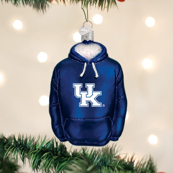 A Christmas tree ornament shaped like a navy blue hoodie with UK in white letters on the front, hanging on a green branch against a background of blurred white lights.