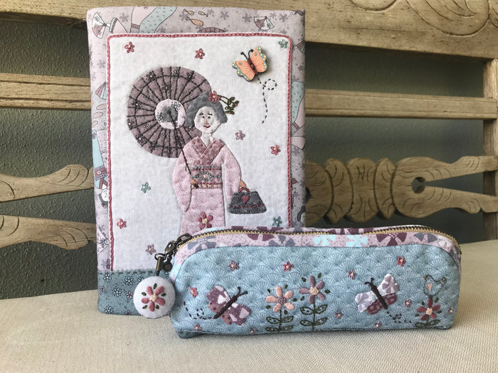 A fabric-covered journal and pencil case set featuring embroidery with a floral and geometric pattern, predominantly in shades of pink, mauve, and blue.
