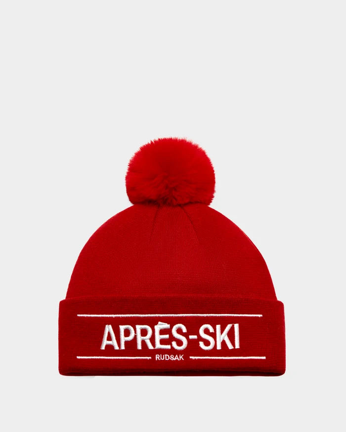 The KRISS - 9424500 Flame Red by Rudsak is a bold unisex ski tuque with a large pom-pom and cuff embroidered in white with APRÈS-SKI above RUDSAK. This classic après-ski essential pops against a plain white background.