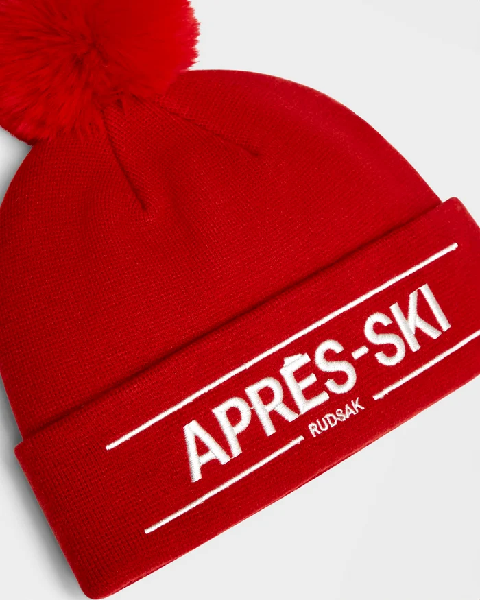 The KRISS - 9424500 Flame Red by Rudsak is a cozy unisex winter hat with a large pom-pom and APRÈS-SKI and RUDSAK embroidered in white on the front fold—perfect for après-ski style.
