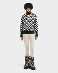 A person wearing Rudsak’s MAGGIE - 8525645 Vanilla Monogram boots, paired with a black fur hat, a women’s black-and-white geometric ski sweater, and cream leggings stands against a white background.