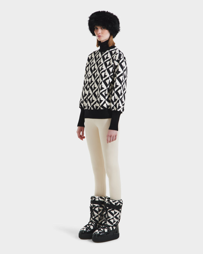 A woman stands against a plain white background wearing the Rudsak MAGGIE - 8525645 Vanilla Monogram ski sweater, styled with white pants, matching winter boots, black gloves, and a fluffy black hat.