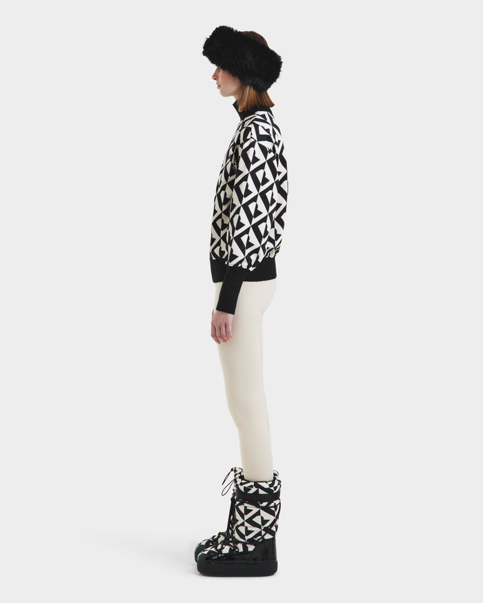 A woman stands in profile wearing Rudsak’s MAGGIE - 8525645 Vanilla Monogram winter boots, paired with a black fur visor, a geometric women’s ski sweater, and white pants against a plain white background.