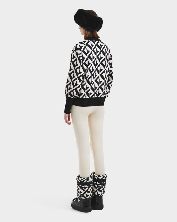 A person stands facing away, wearing the Rudsak MAGGIE - 8525645 Vanilla Monogram women’s ski sweater, stylish boots, white pants, and a black furry headband against a plain white background.