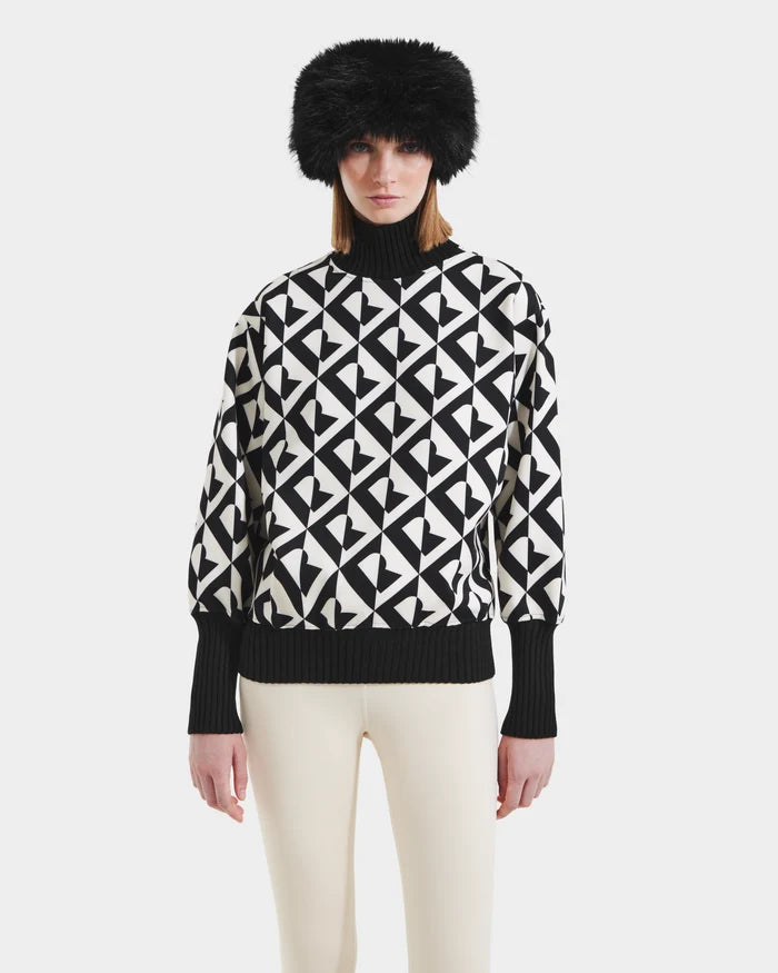 A woman models the Rudsak MAGGIE - 8525645 Vanilla Monogram ski sweater with black-and-white geometric patterns, paired with a black furry hat and white pants, standing against a plain white background.