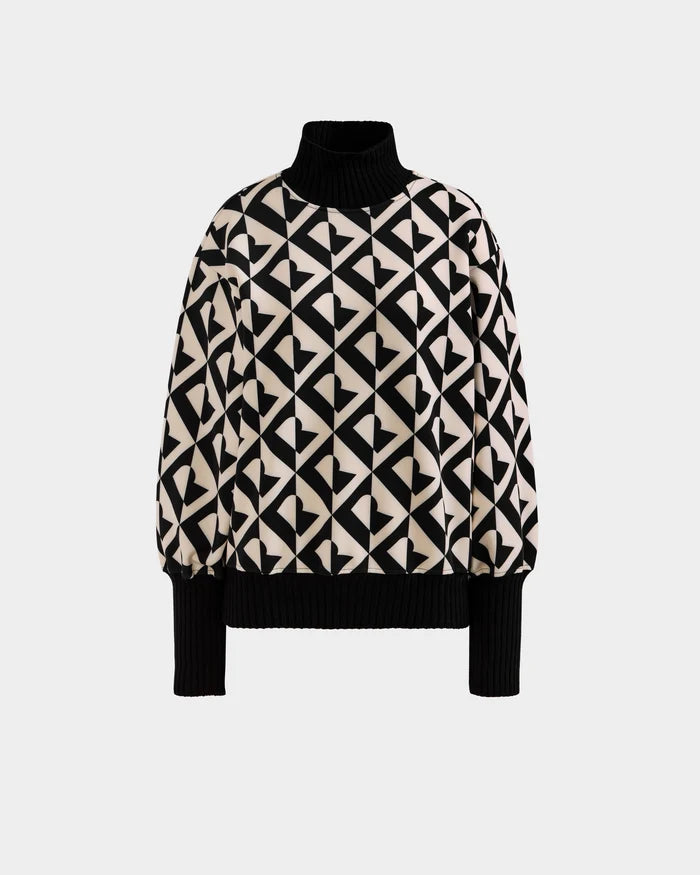 The Rudsak MAGGIE - 8525645 Vanilla Monogram is a women’s long-sleeve ski sweater featuring a black and white geometric heart pattern with ribbed black turtleneck, cuffs, and hem, shown on a plain white background.