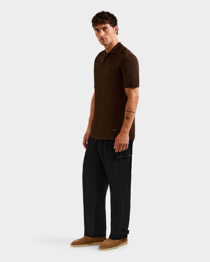 A man faces forward wearing the Rudsak MAISEN - 6524518 Mocha knit polo, paired with loose black pants and tan shoes, against a plain light gray background.