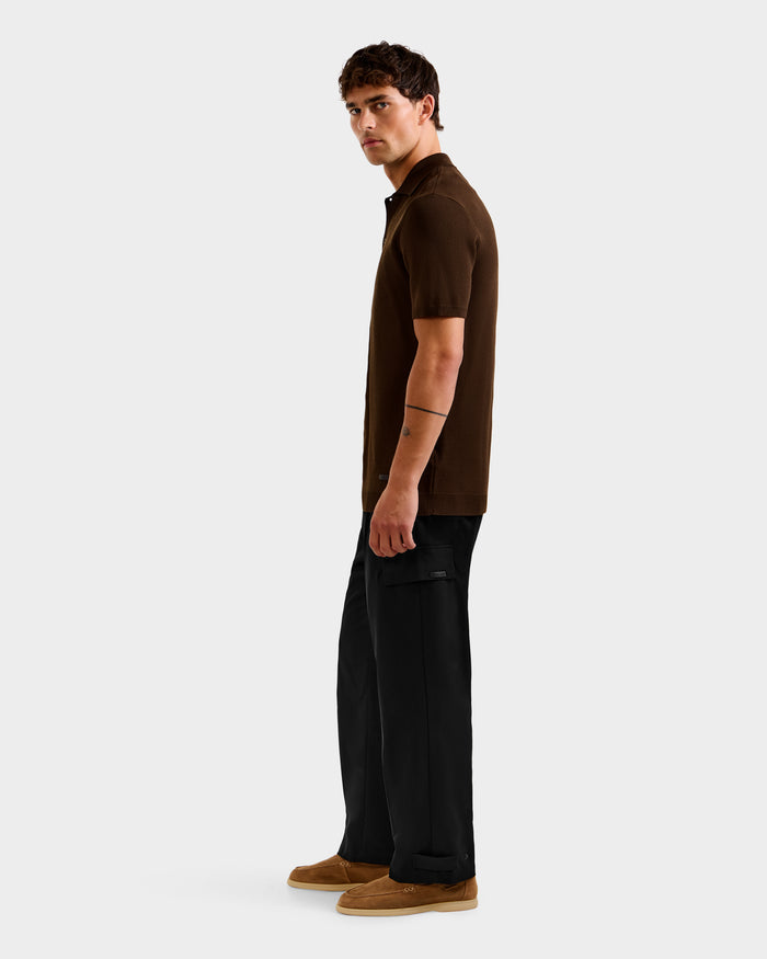 A man with short dark hair and a visible arm tattoo stands sideways against a plain background, wearing the Rudsak MAISEN - 6524518 Mocha short-sleeve polo shirt, black cargo pants, and tan shoes.