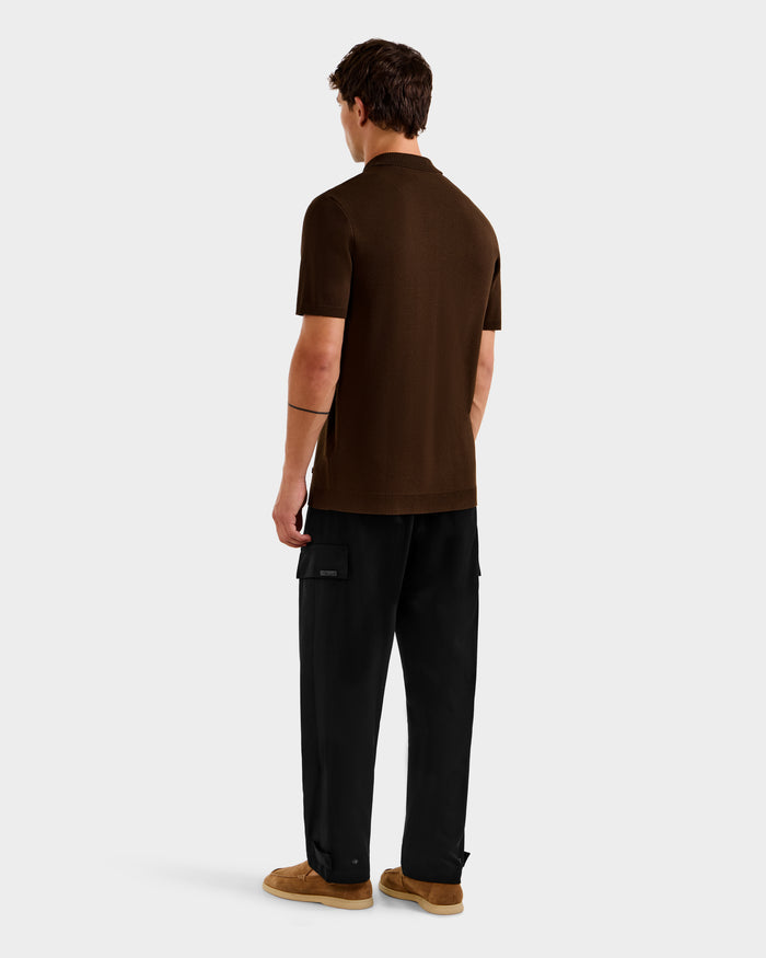 A person stands facing away, wearing the Rudsak MAISEN - 6524518 Mocha short-sleeve men's knit polo with black cargo pants and tan shoes against a light background—a modern men's wardrobe staple.