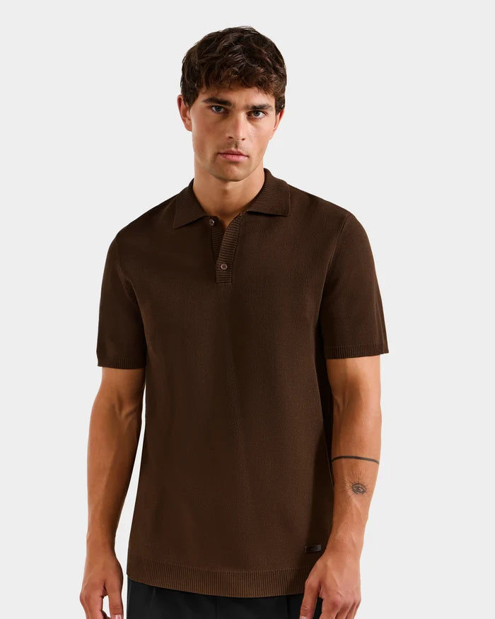 A man with short dark hair wears the Rudsak MAISEN - 6524518 Mocha men's knit polo and black pants, standing against a plain light gray background—a timeless addition to the modern wardrobe.