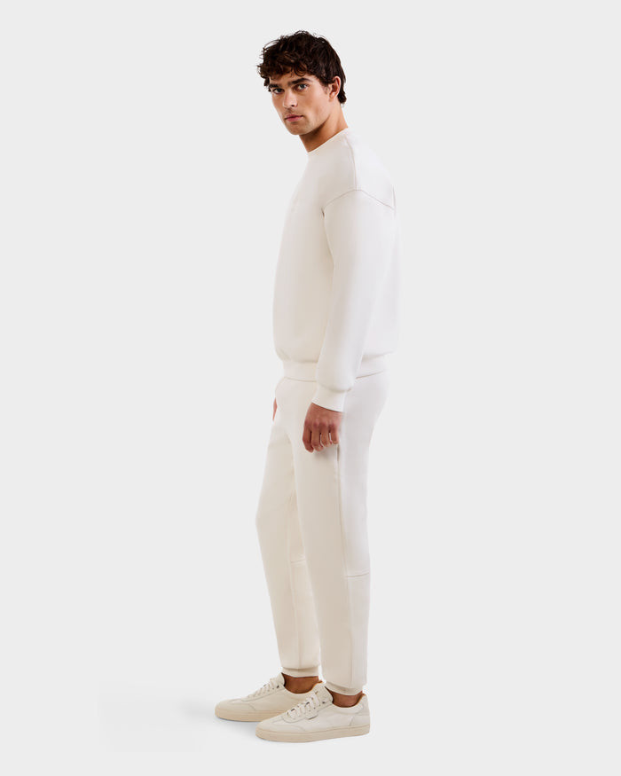 A man models the Rudsak MAXTON - U525007 Vanilla M jogger and sweatshirt set with white sneakers, showcasing casual comfort in an all-white look against a plain backdrop.