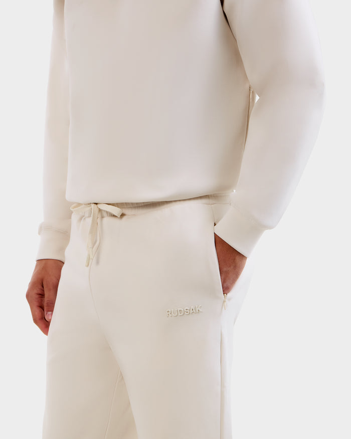 A person wears the Rudsak MAXTON - U525007 Vanilla M jogger sweatshirt and sweatpants set, featuring cream-colored cotton-blend fabric and embroidered "RUDSAK" near the pocket for everyday comfort.