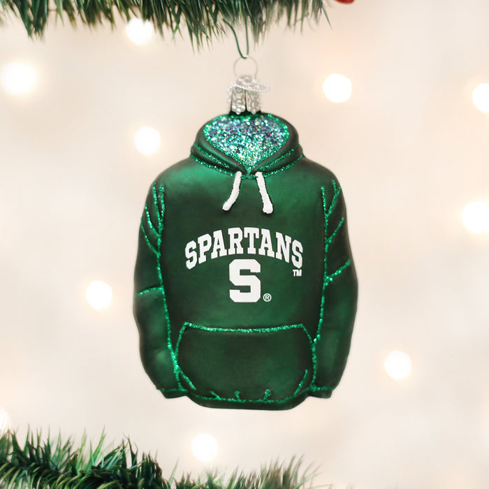 A green hoodie-shaped Christmas ornament with SPARTANS and an S logo on the front, hanging on a tree branch. The ornament has glitter accents and a festive background with soft lights.