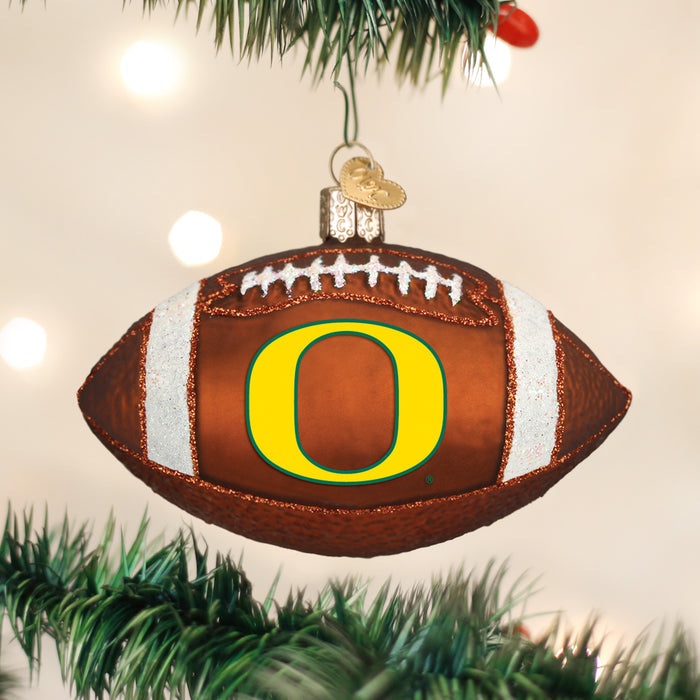A football-shaped Christmas ornament with the Oregon Ducks O logo hangs on a Christmas tree, surrounded by green branches and soft white lights in the background.
