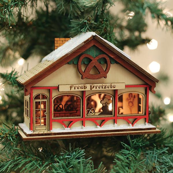 A wooden ornament shaped like a bakery labeled Fresh Pretzel with pretzel decorations and snow on the roof, hangs on a Christmas tree with blurred lights in the background.