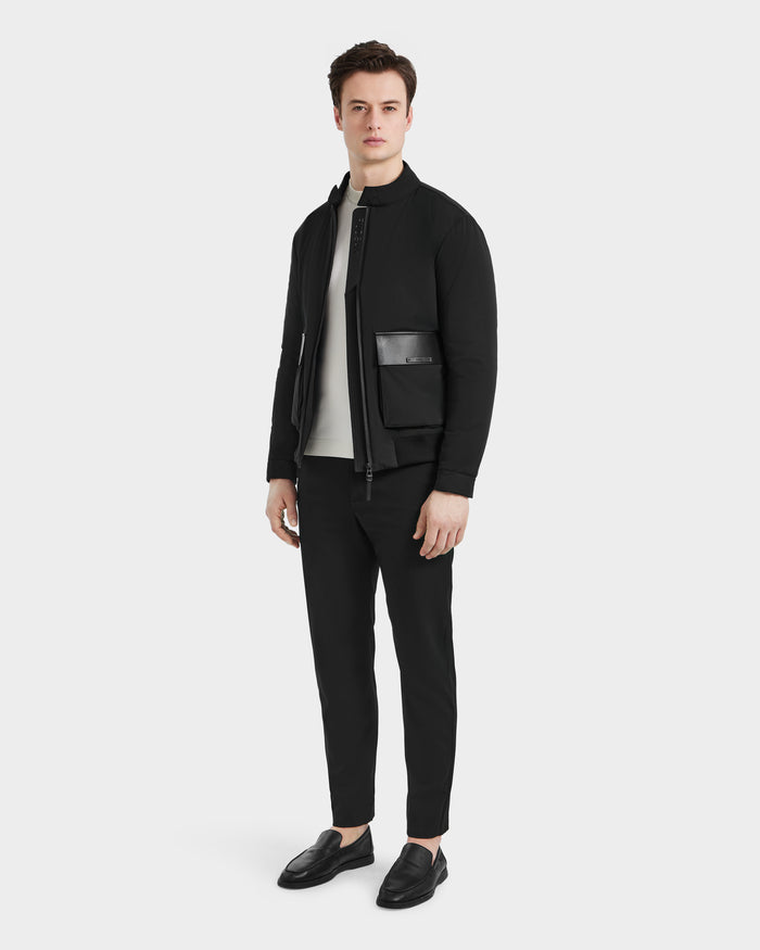A man stands against a plain white background, wearing the Rudsak PETER - 6125001 Black bomber jacket with vegan leather accents, a light gray shirt, black pants, and black loafers. He looks forward with a neutral expression.