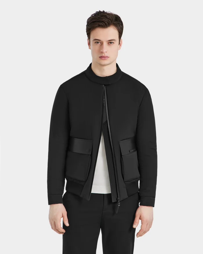 A young man with short brown hair models the Rudsak PETER - 6125001 Black zip-up bomber jacket with vegan leather accents, styled over a white shirt and black pants, standing against a light gray background with a neutral expression.