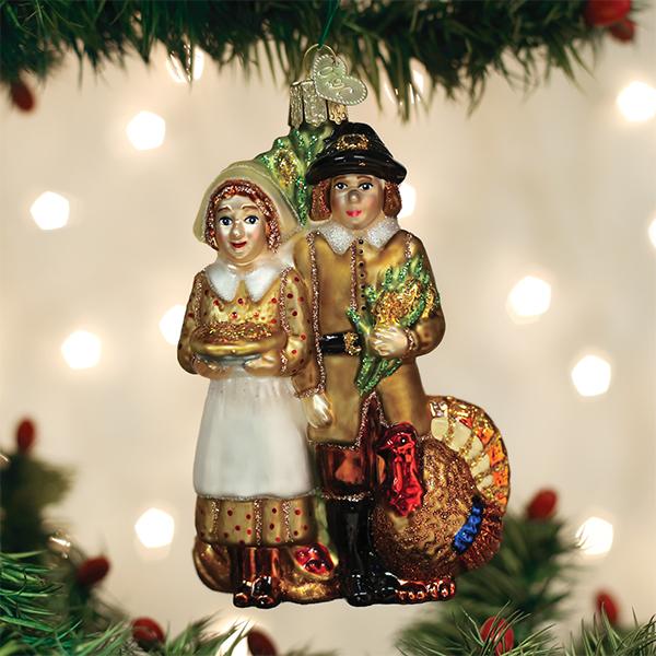 A Christmas tree ornament depicting a Pilgrim couple in traditional attire, with the woman holding a pie and the man standing next to a turkey, hangs among green branches with twinkling lights in the background.