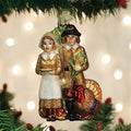A Christmas tree ornament depicting a Pilgrim couple in traditional attire, with the woman holding a pie and the man standing next to a turkey, hangs among green branches with twinkling lights in the background.