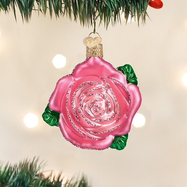 A pink rose-shaped ornament with glitter accents hangs on a Christmas tree branch with blurred white lights in the background.