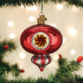 A shiny red Christmas ornament with gold and yellow accents hangs on a green Christmas tree branch, with blurred lights glowing softly in the background.